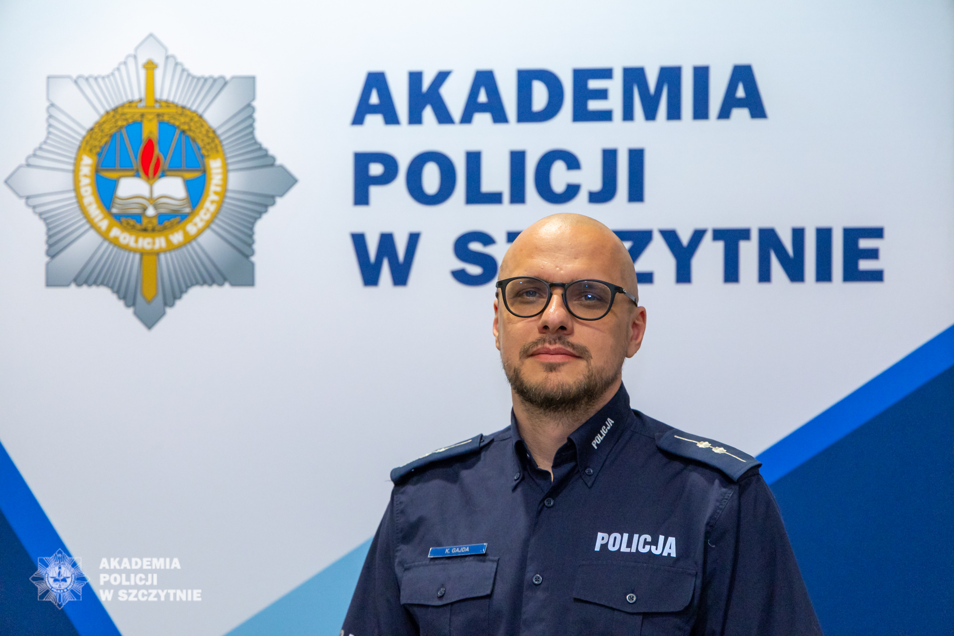 Aspirant Karol Gajda stoi w umundurowaniu służbowym na tle ścianki Akademii Policji w Szczytnie.
